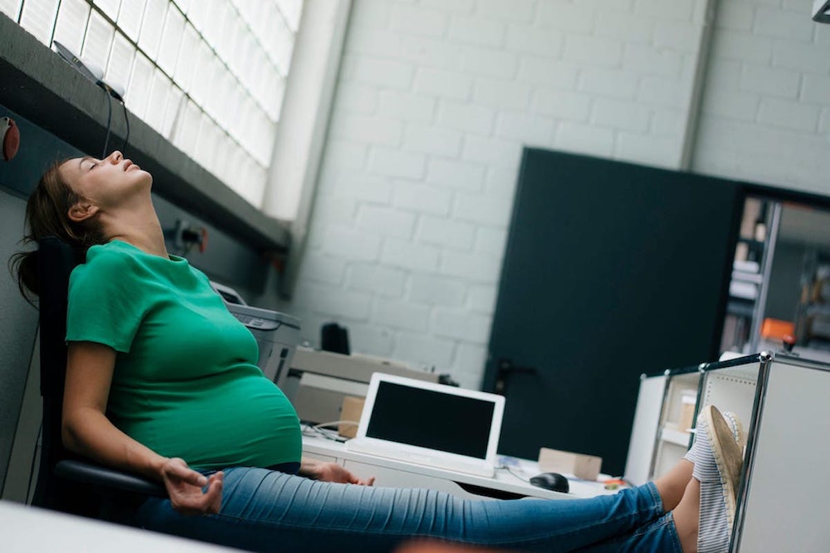 A heavily pregnant person with her feet up at her office closes her eyes.