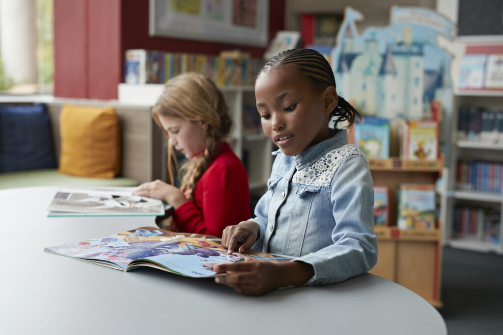 Two young students sit at a table in the library, paging through books.