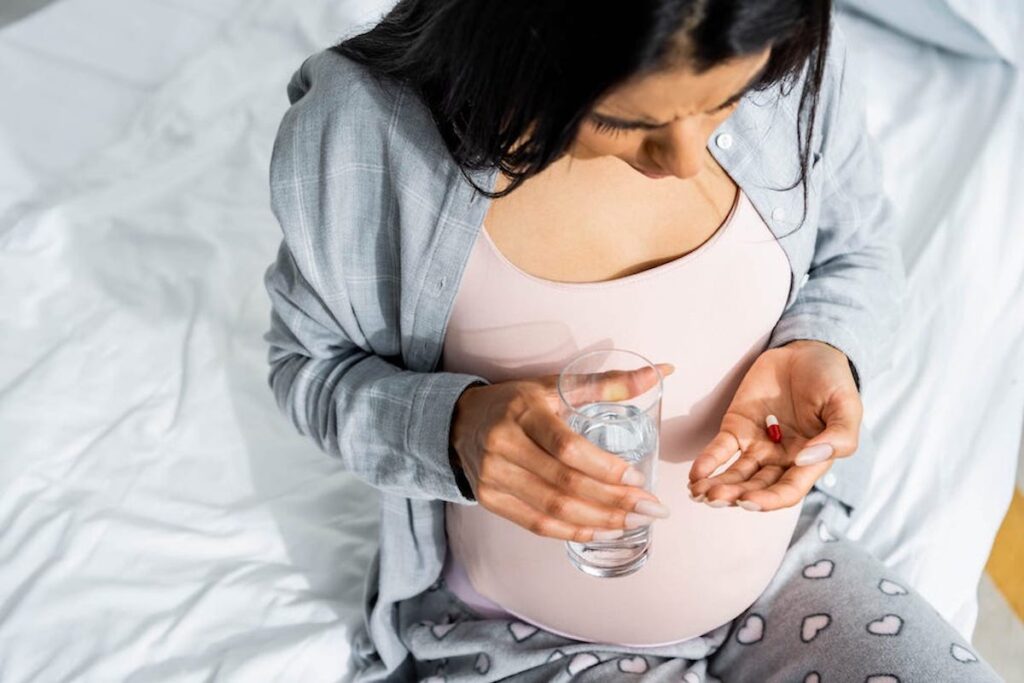 A pregnant person sits on a bed, holding a glass of water and a pill. She is looking down at the pill.