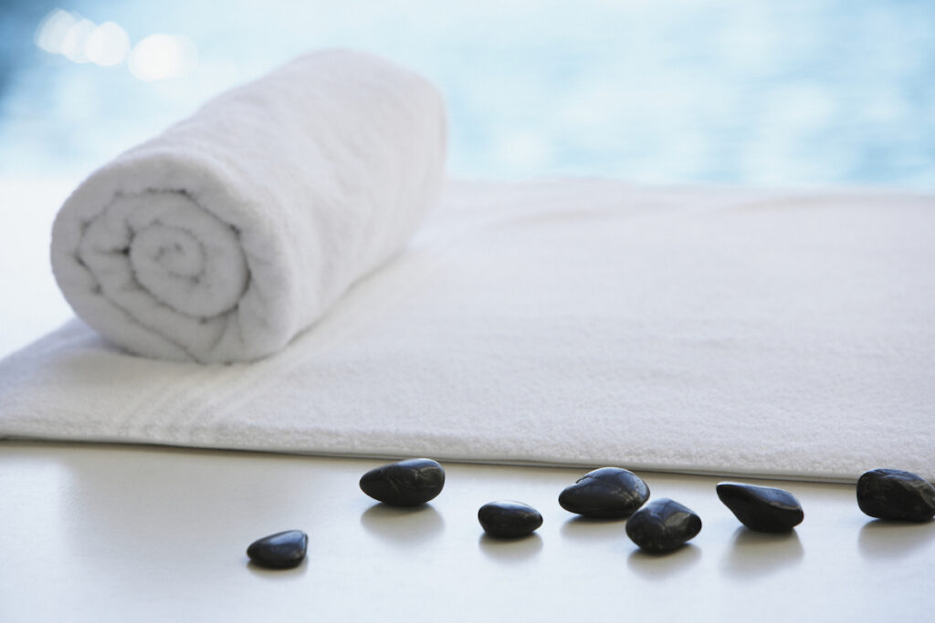 A rolled up white towel and massage stones are seen in front of a blue pool.