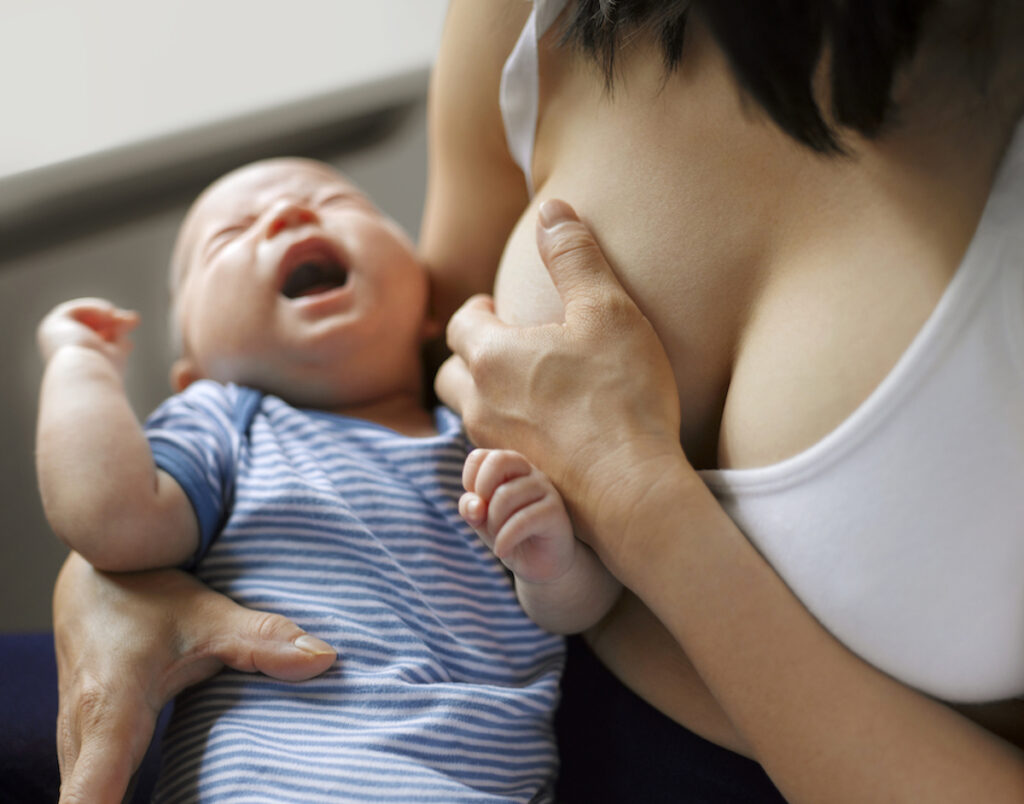 A baby cries while a breastfeeding woman suffering from mastitis cups her breast in pain.