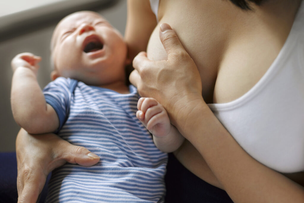 A baby cries while a breastfeeding woman suffering from mastitis cups her breast in pain.