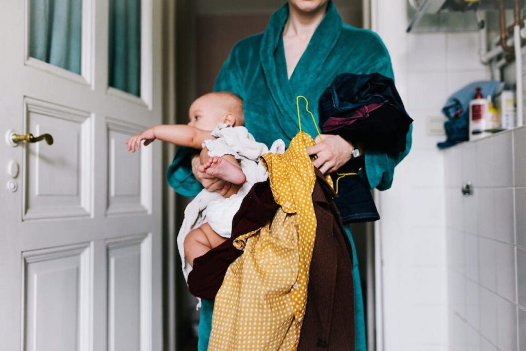 A woman stands at the door in a green bathroom. She is holding a baby in one arm and hangers with different clothing items in the other.