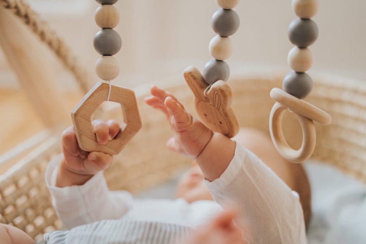 A baby reaches out of a basket to grab wooden rings on a mobile.