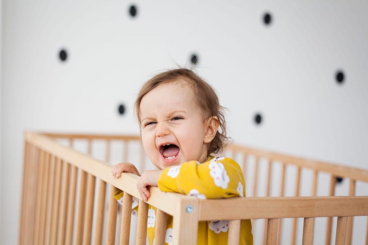 A baby wearing a yellow onesie screams in a crib in front of black and white polkadot wallpaper.