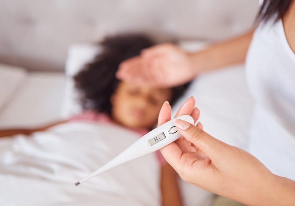A parent holds the back of her palm to her child's forehead while the child sleeps in bed. In the other hand, the parent holds a thermometer that reads "hi"