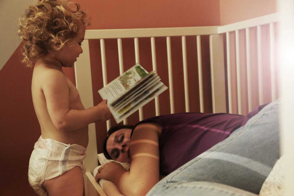 A parent sleeps in a toddler bed while the toddler stands over the parent holding a book.