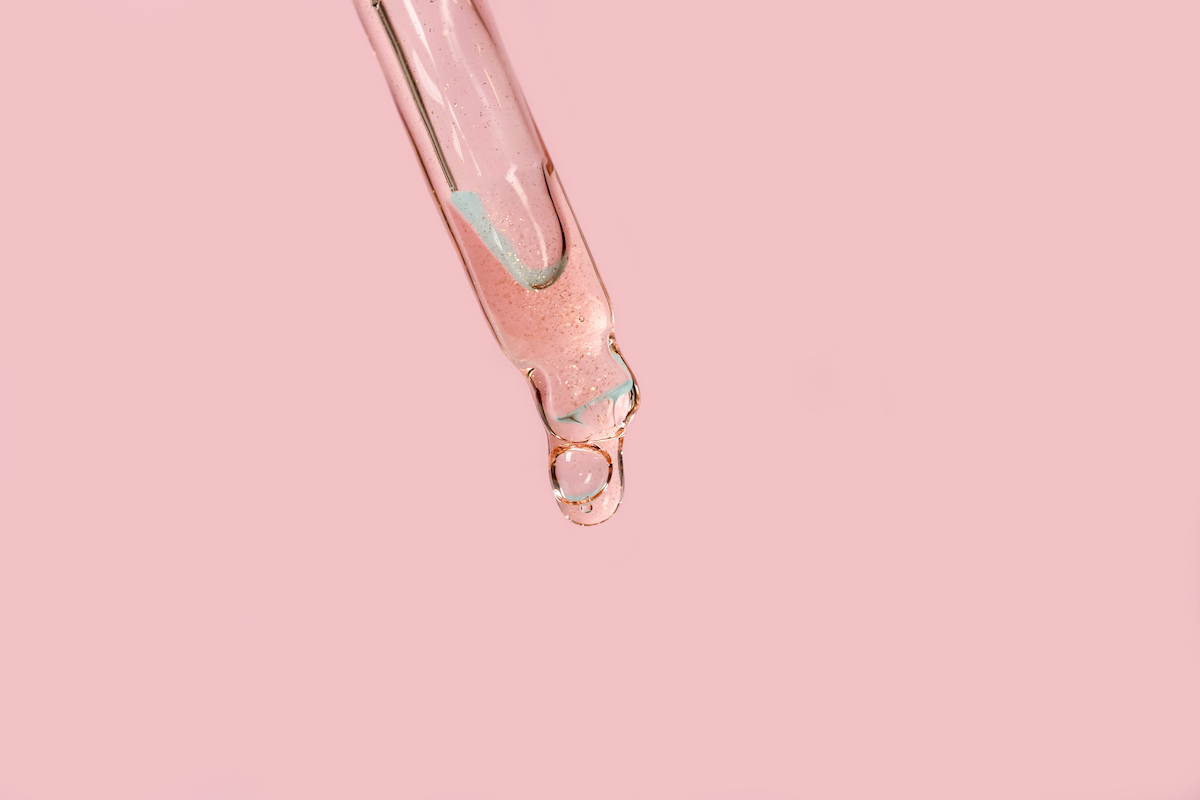Glass pipette dripping oil in front of a pink background.