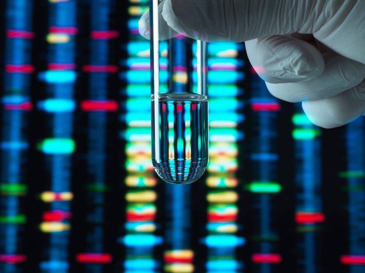 A gloved hand holding a glass test tube with clear liquid is seen in from of a glowing screen showing DNA comparisons.