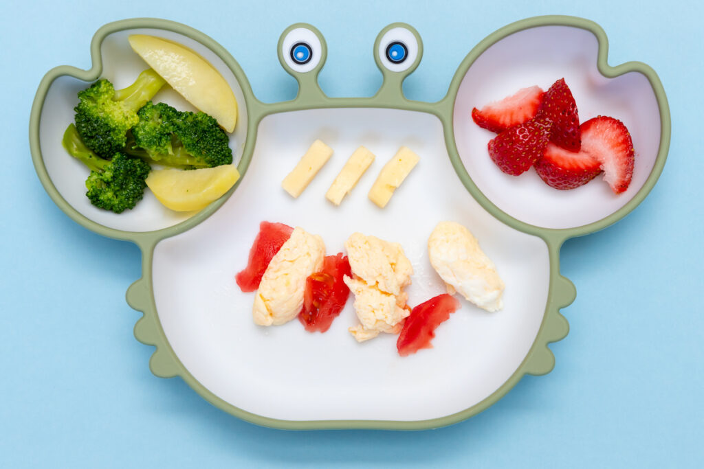 An above view of a toddler plate shaped like a crab and filled with broccoli, chicken, and strawberries.