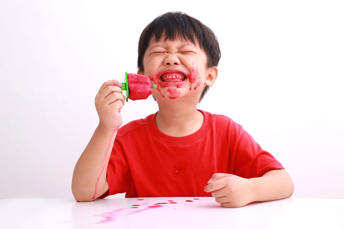 A kid in a red shirt smiles while eating a red popsicle that has dripped all over their face and arm.