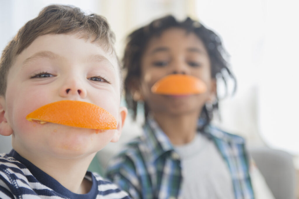 Two kids bite orange slices to cover their smiles with the peel.