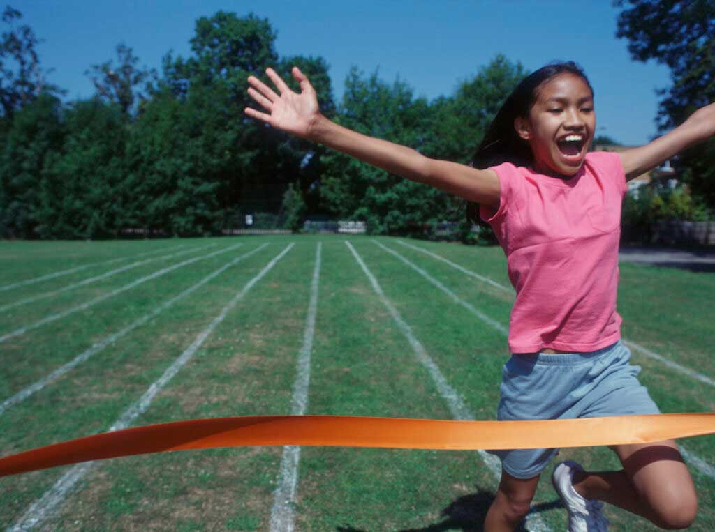 A girl, with arms outstretched, crosses the finish line on a green field.