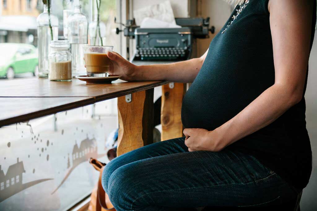 Person with a pregnant belly sits by the window drinking coffee in a cafe.