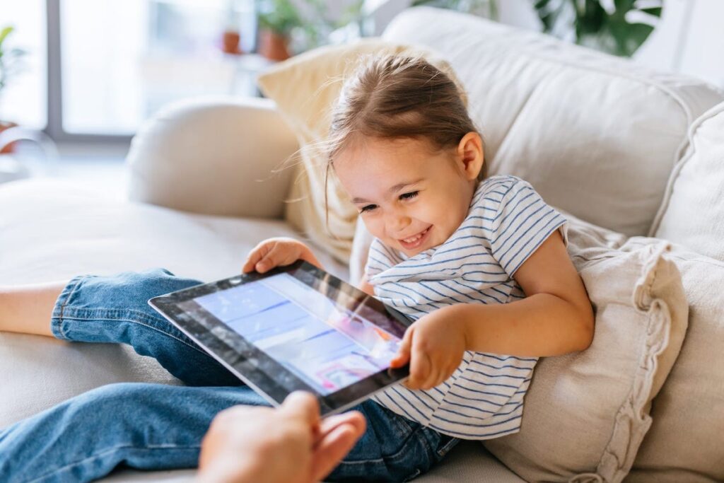 A child sits on a couch playing with an ipad that she won't hand over to her parent.