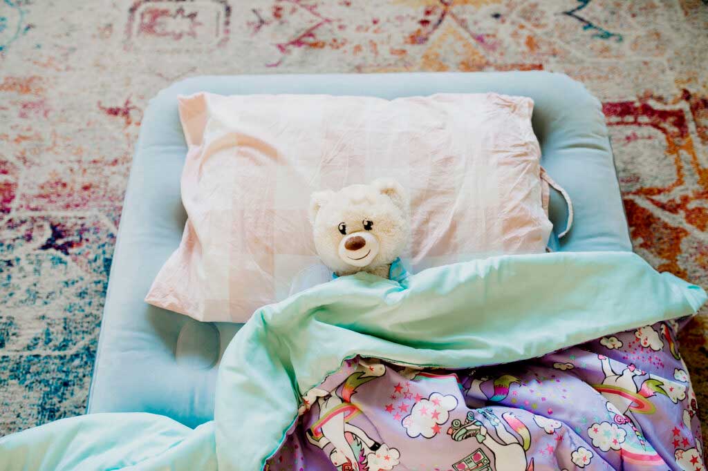 A blue air mattress is set up on a carpeted floor with a teddy bear and unicorn blanket.