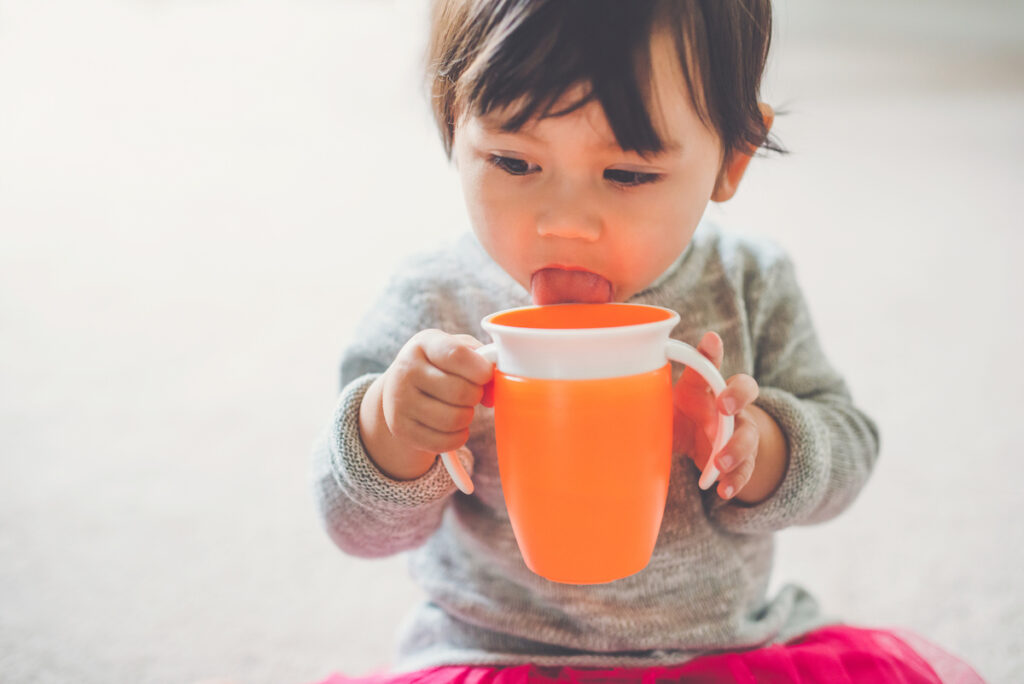A small toddler holds and orange sippy cup and licks the top of it.