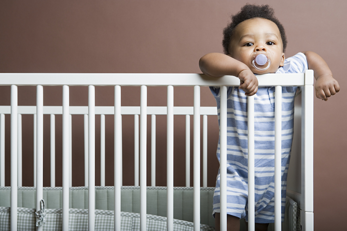 An infant with a pacifier stands inside a crib, holding onto the rails and looking at the camera.