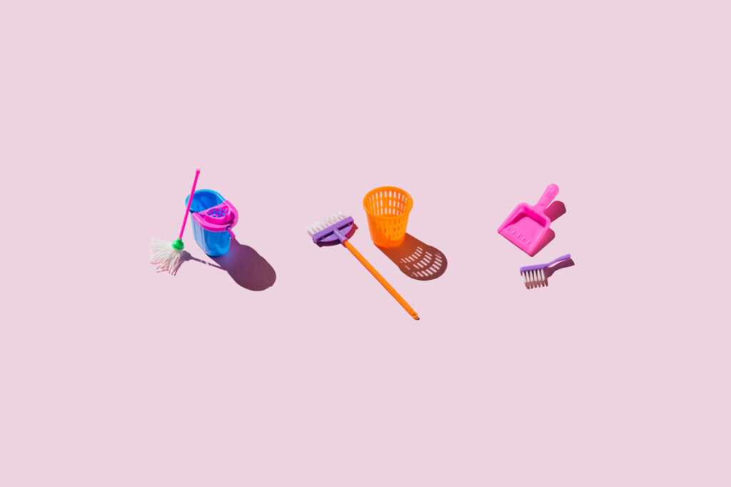Toy-sized mop and bucket, broom and garbage can, and brush and dustpan are seen on a pink background.