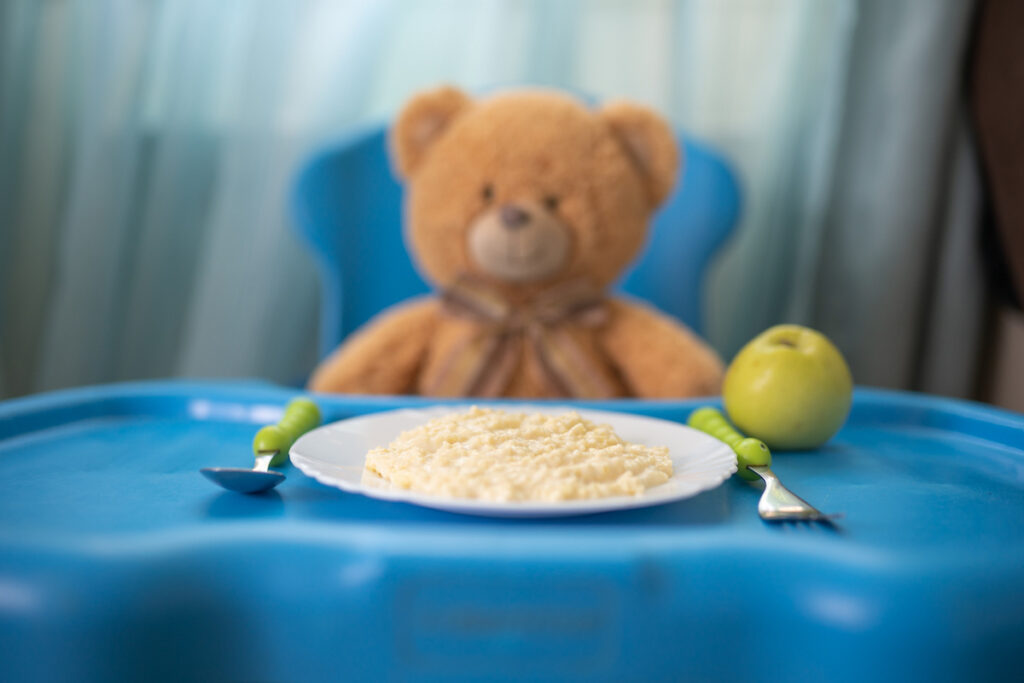 A teddy bear sits in a blue highchair eating oatmeal and an apple.