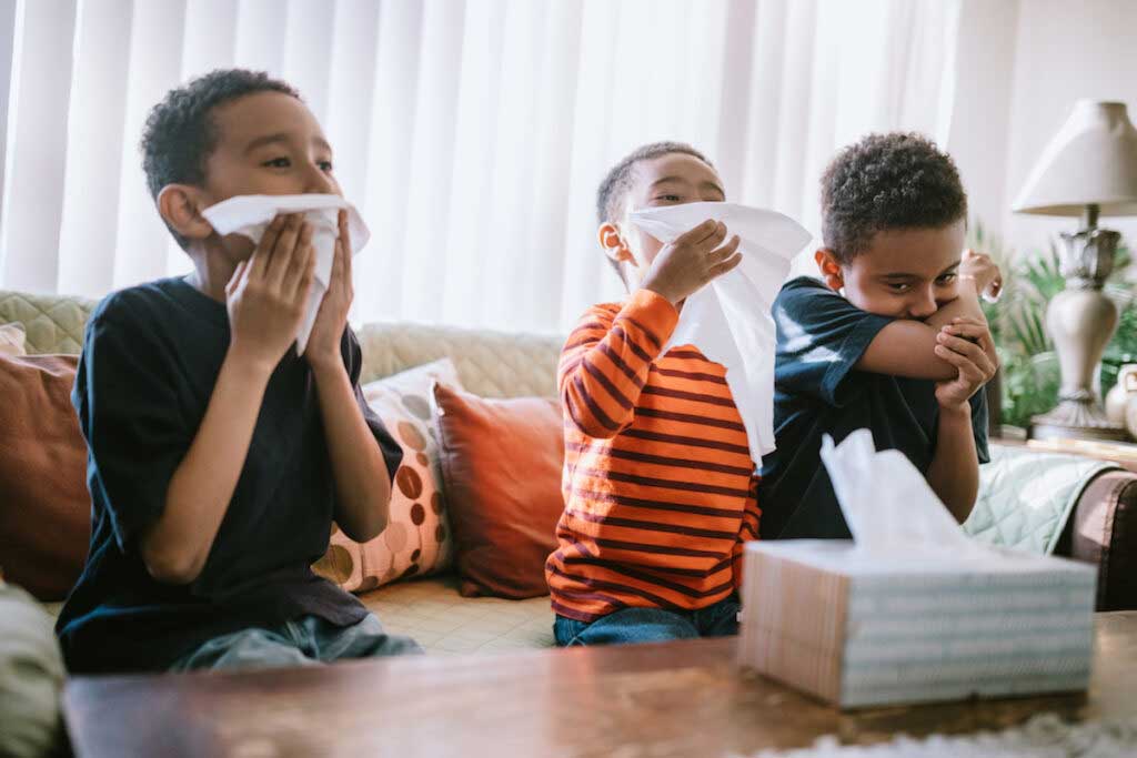 Three kids in a family sit on a couch, each holding a tissue paper to their nose.