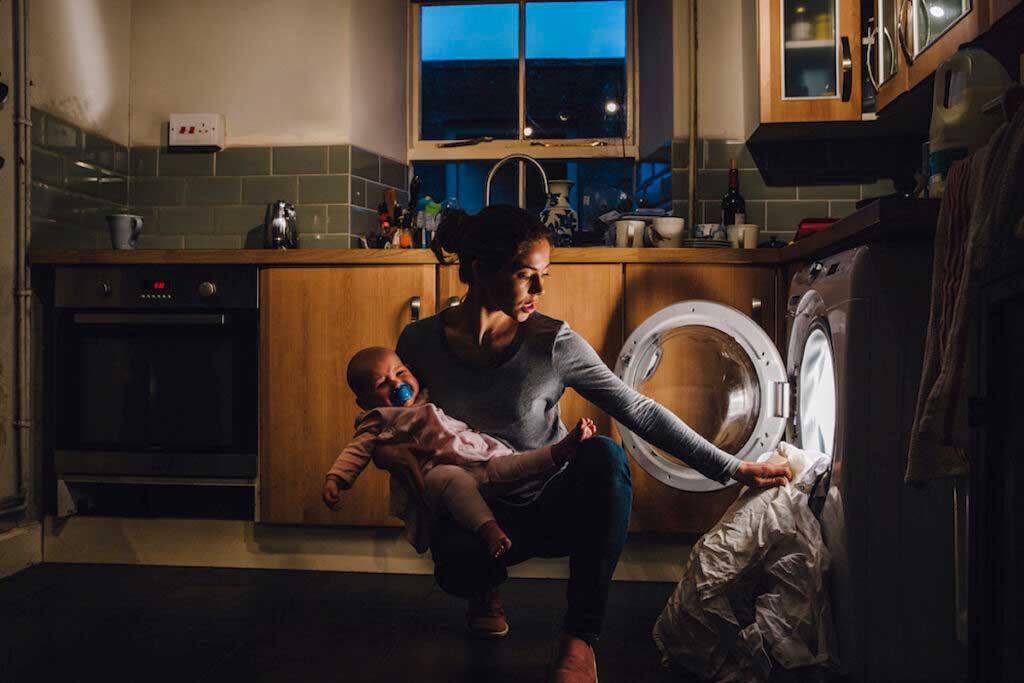 A young parent is holding a crying baby on one hip while trying to do the laundry with her other hand.