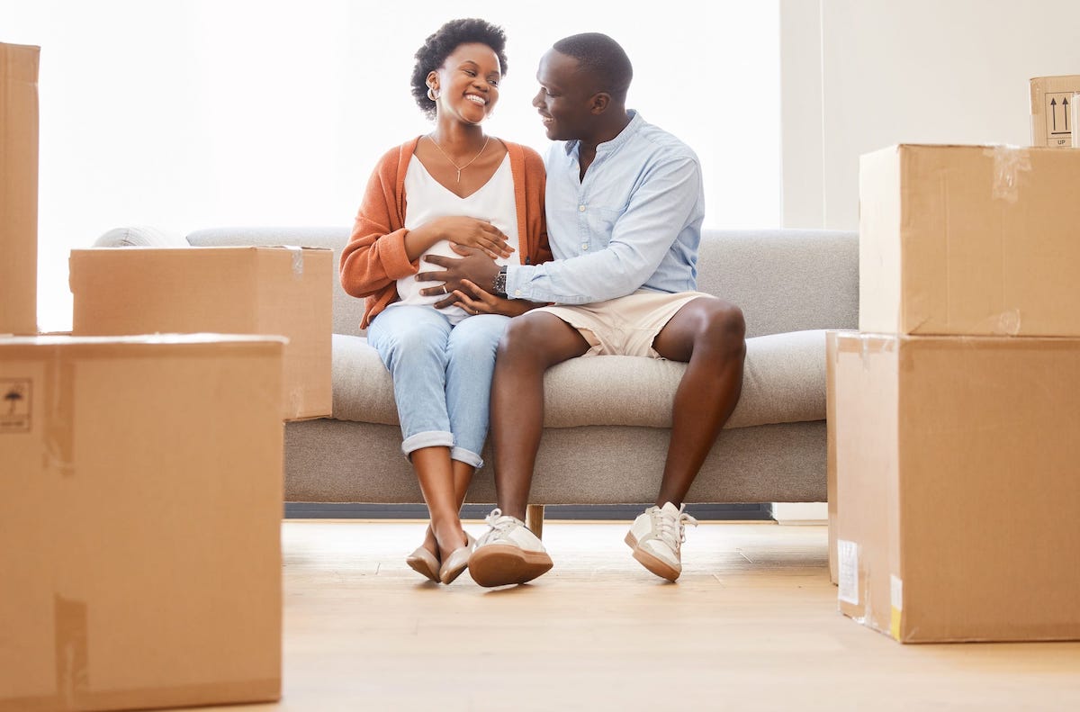 A pregnant person and partner sit on a couch surrounded by brown boxes of baby items.