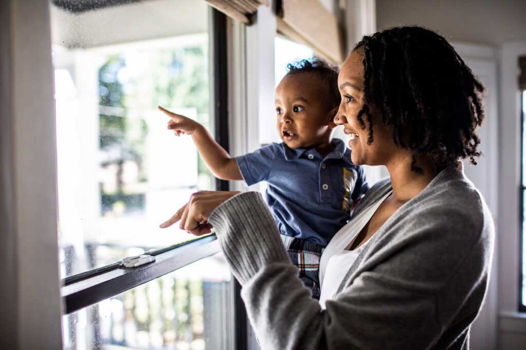 Parent holds a young toddler while looking at the window. They are both pointing to objects outside.