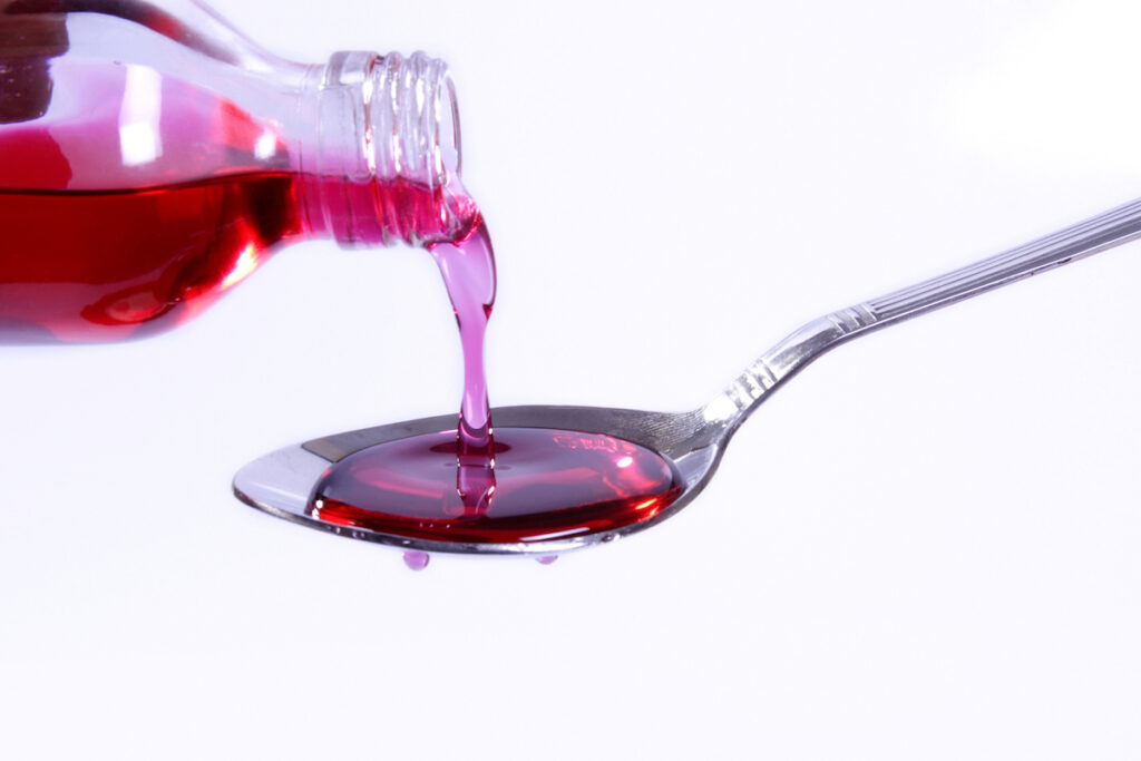 Cherry-red cough syrup is poured from a bottle onto a dripping spoon.