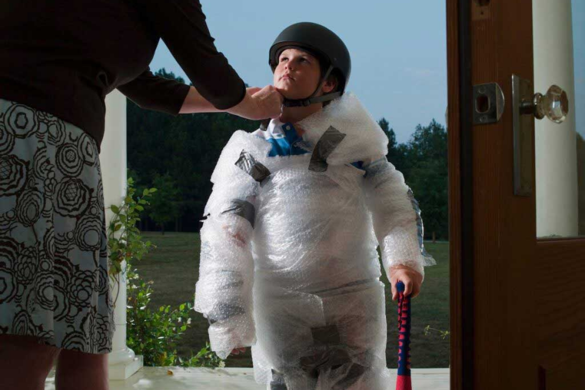 A child prepares to go out to play by getting wrapped in bubble wrap and wearing a helmet.