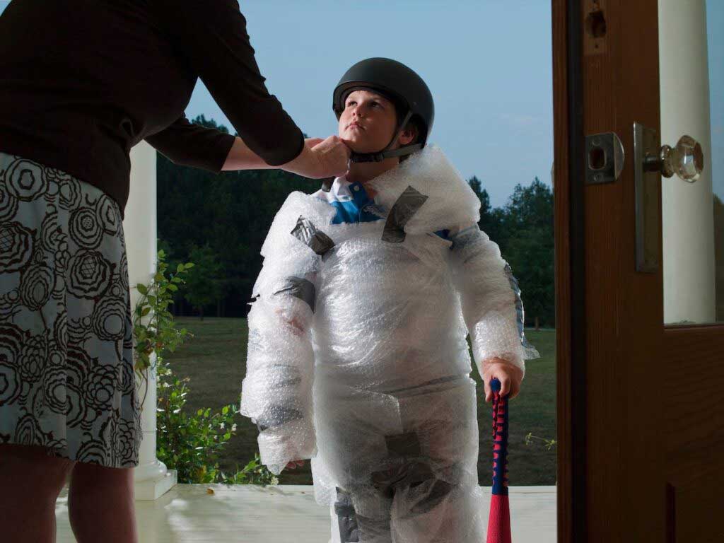 A child prepares to go out to play by getting wrapped in bubble wrap and wearing a helmet.