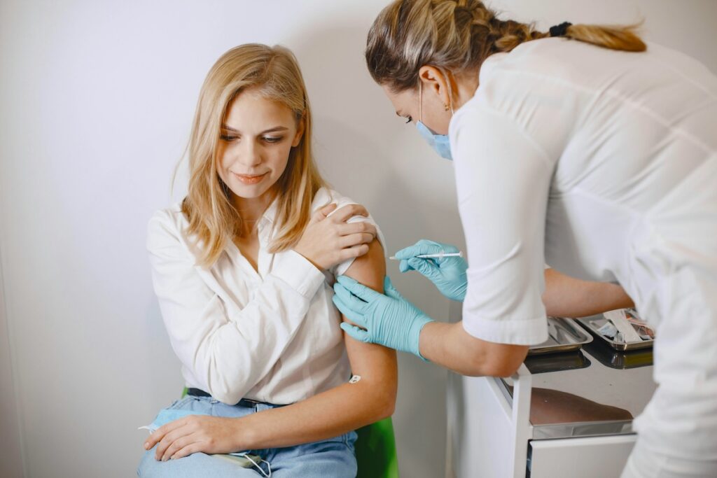 Blonde woman getting a vaccine from masked blonde doctor.