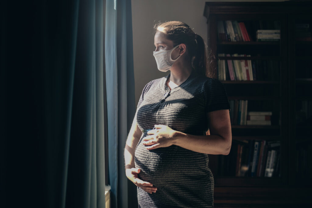 A pregnant person in a hazy room stands by a window wearing a mask.