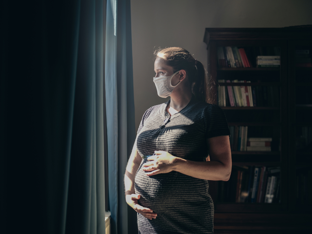 A pregnant person in a hazy room stands by a window wearing a mask.