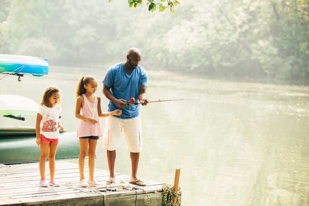 Parent and two children stand on a dock over a pond on a muggy summer day while the parent fishes.