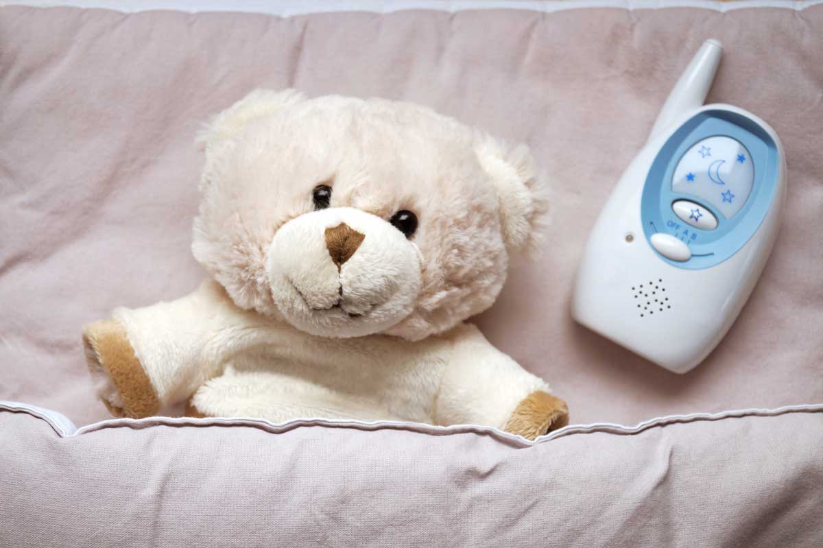 A teddy bear is tucked under a pink sheet with an audio-only baby monitor.