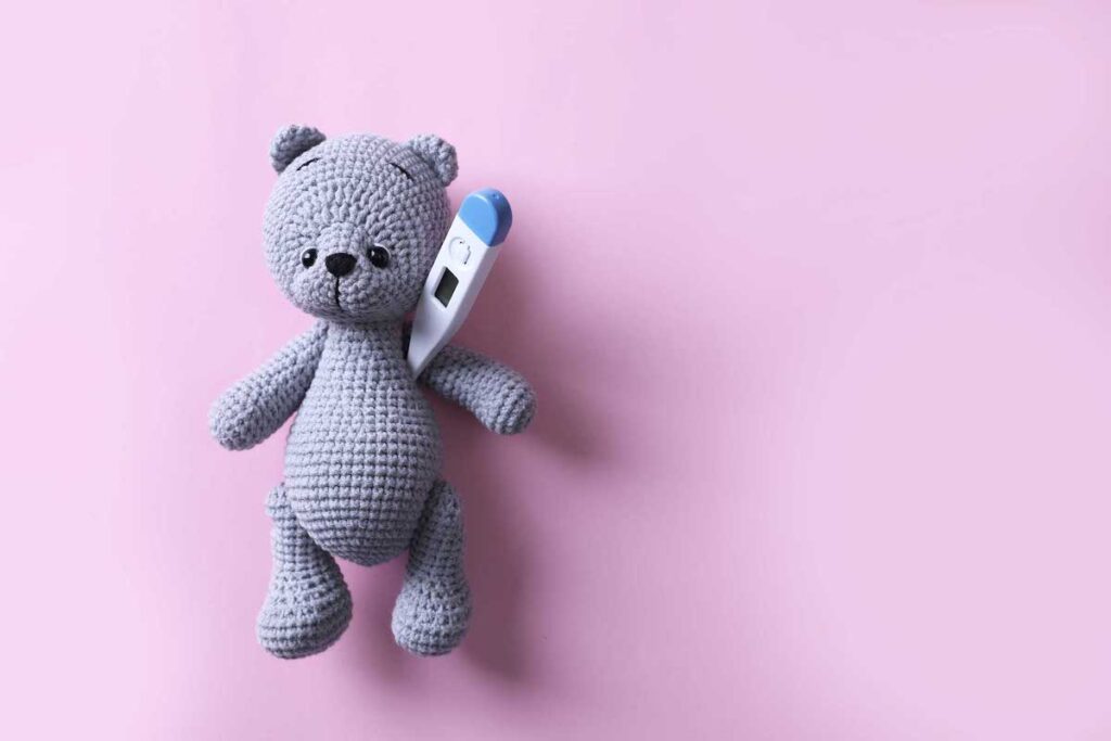 A gray knit bear on a pink background has a thermometer under its arm.