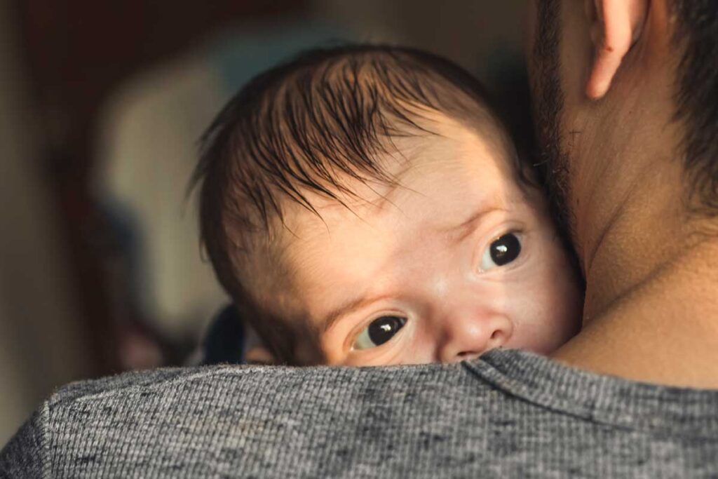 An over-the-shoulder shot of a father in a gray t-shirt holding a newborn baby.
