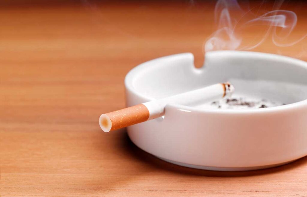 A lit cigarette smokes in an ashtray on a wooden table.