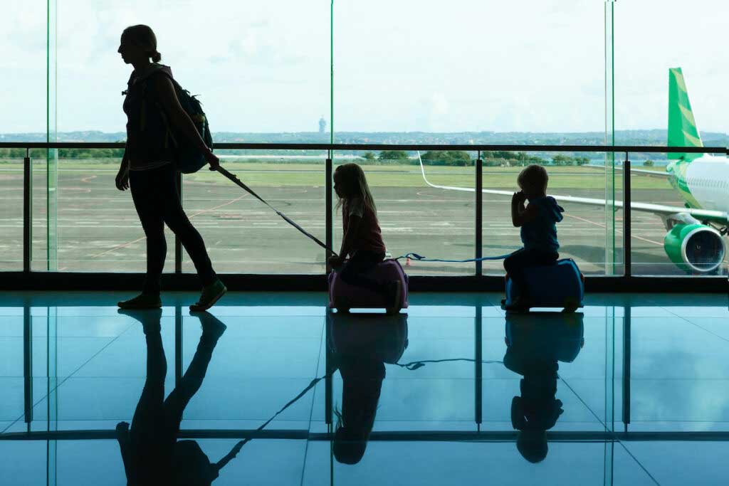In the airport transit hall, a parent pulls two children along as they ride on rolling luggage in front of a large window with an airplane outside.