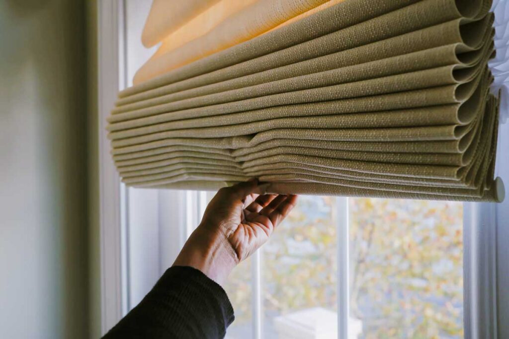 A hand is seen lowering the blinds in front of a closed window on a hazy day.