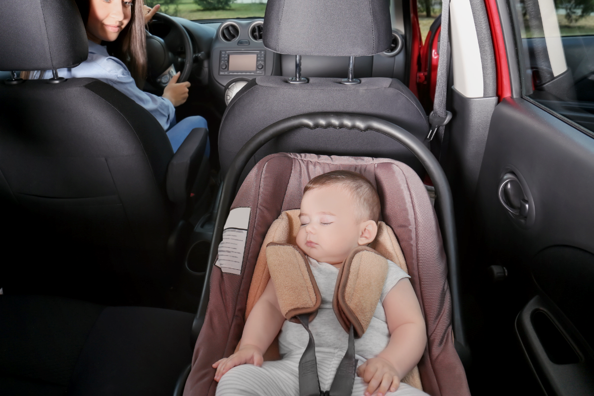 Sleeping baby strapped in a car seat, with a mother looking back and driving.