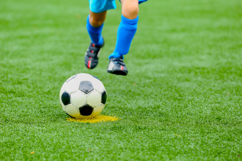 Child's legs, in protective soccer gear, about to kick a soccer ball on artificial turf.