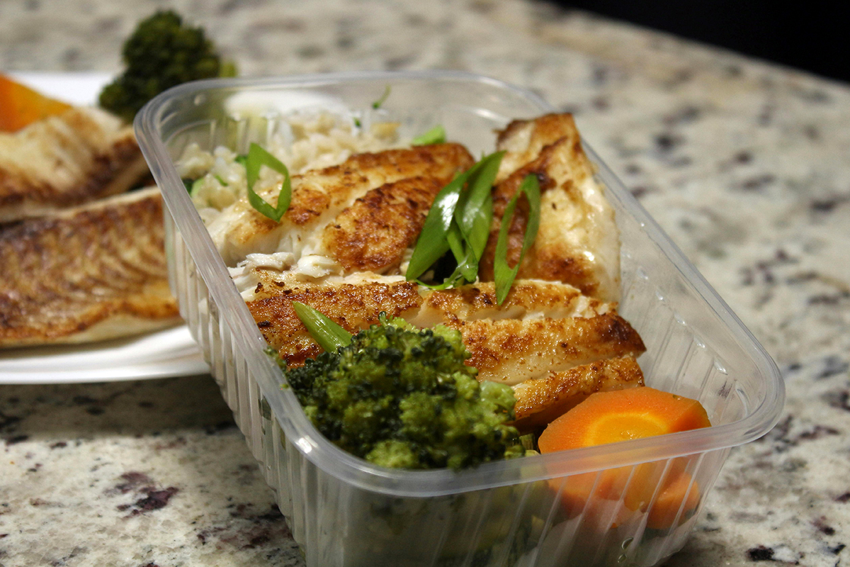 Dinner plate and leftover container with fish, broccoli and carrots on a counter top.