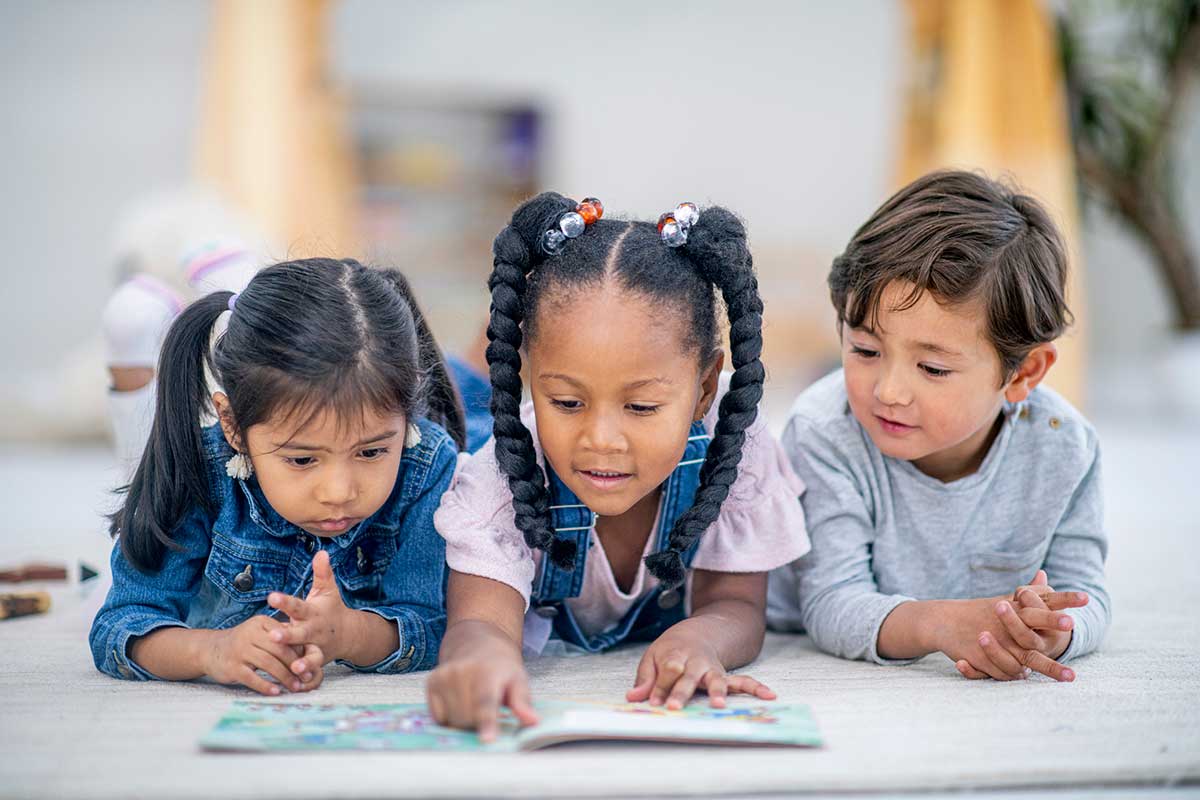 Three children of different races read a book together in a classroom.