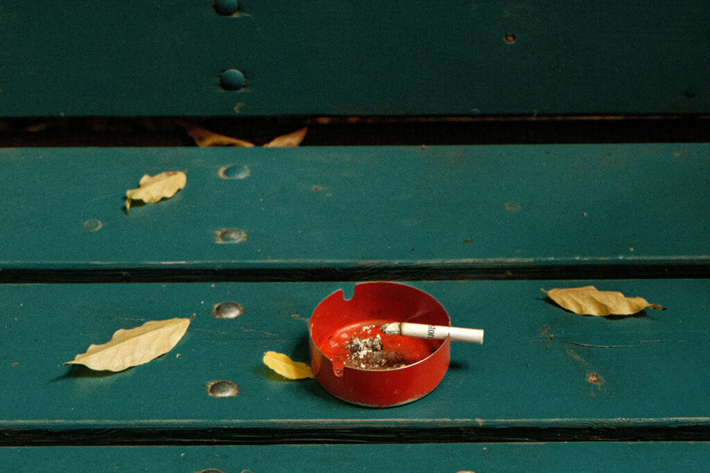 Lit cigarette in a red ashtray on a green bench with fallen leaves around it.