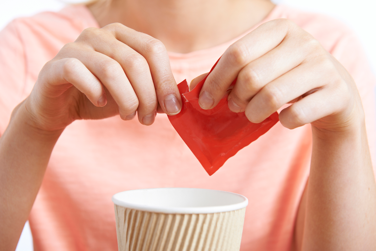 A close up of a person opening a red packet of artificial sweetener over a cup of coffee.