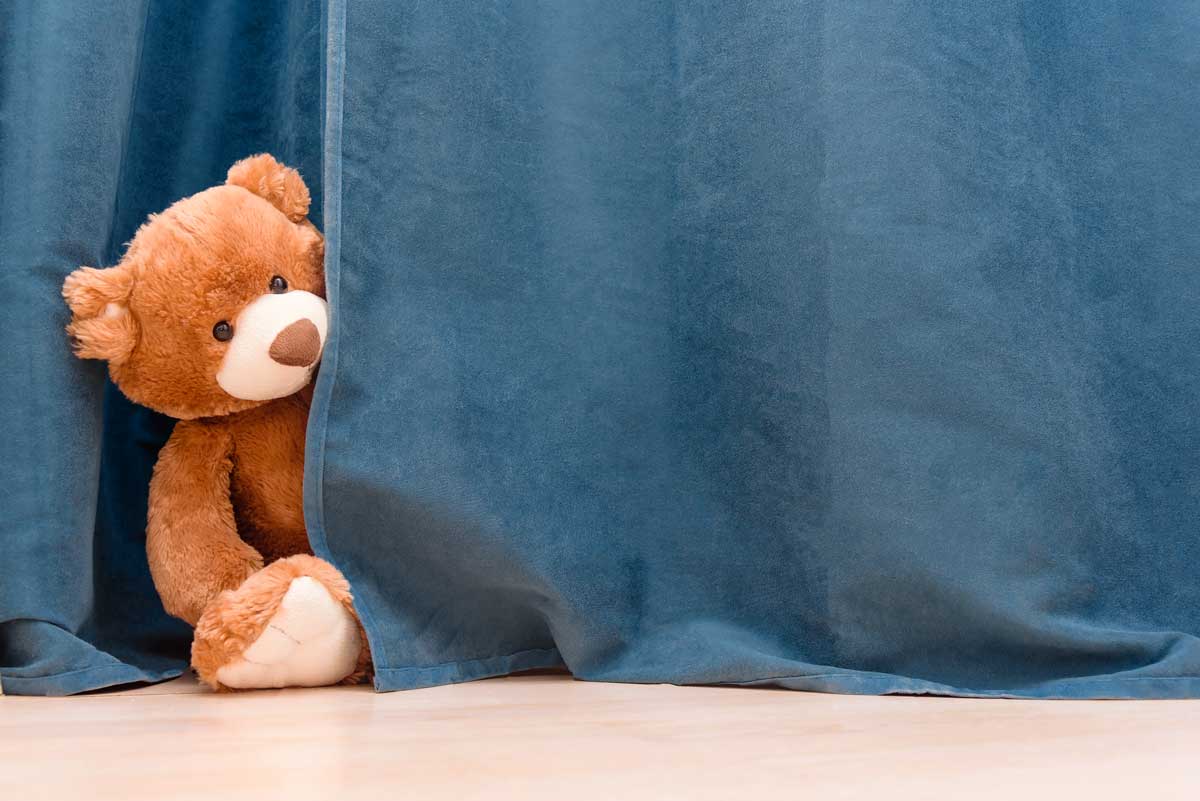 A teddy bear peaks around navy blue blackout curtains.