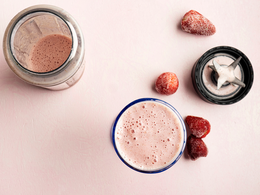 Frozen strawberries, a blender, and cup with a pink smoothie are arranged on a pink background.
