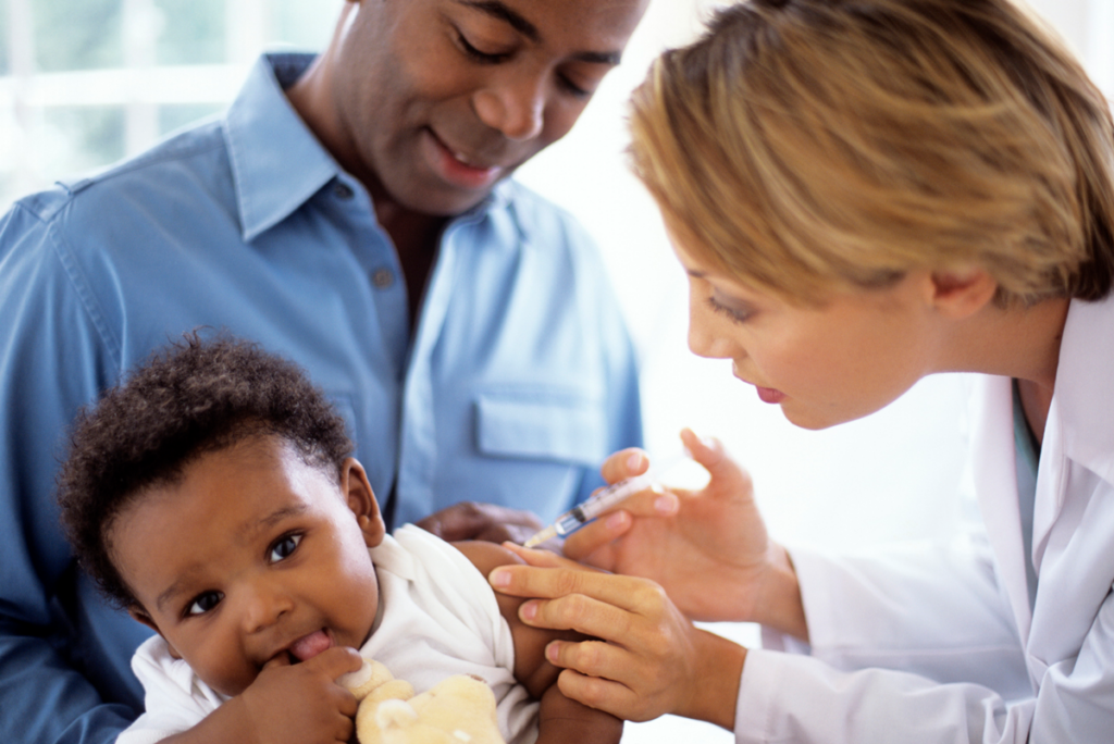 A parent looks on as a pediatrician gives a smiling baby a vaccination.
